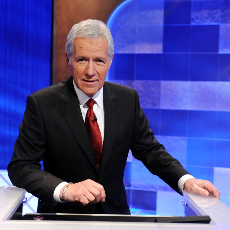 Jeopardy host Alex Trebek stands in front of the Jeopardy board.