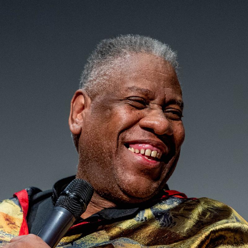 Fashion icon and journalist Andre Leon Talley smiles with a microphone while wearing a brightly colored ensemble 