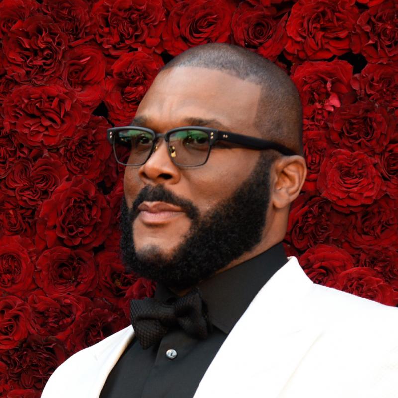 Filmmaker Tyler Perry stands in front of a wall of red roses in a white suit