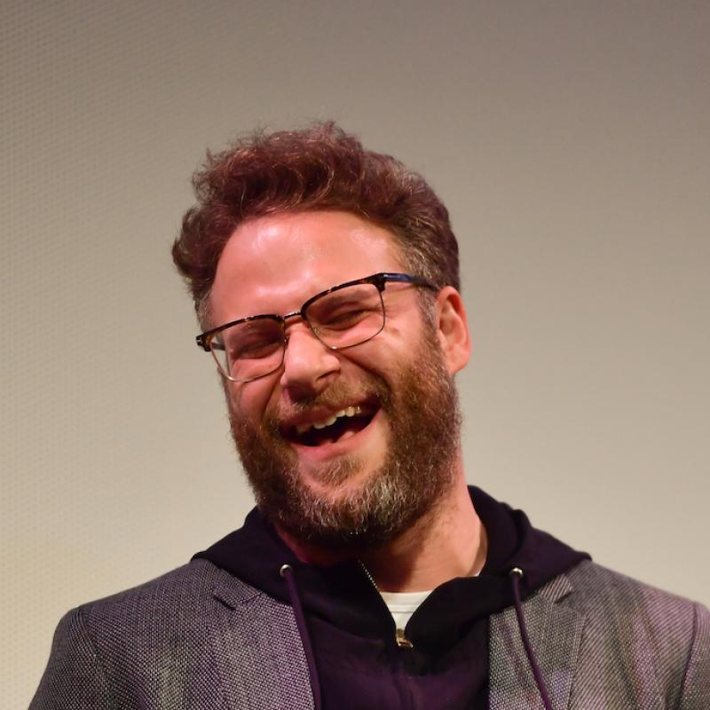 Actor Seth Rogen laughs on stage against a gray backdrop