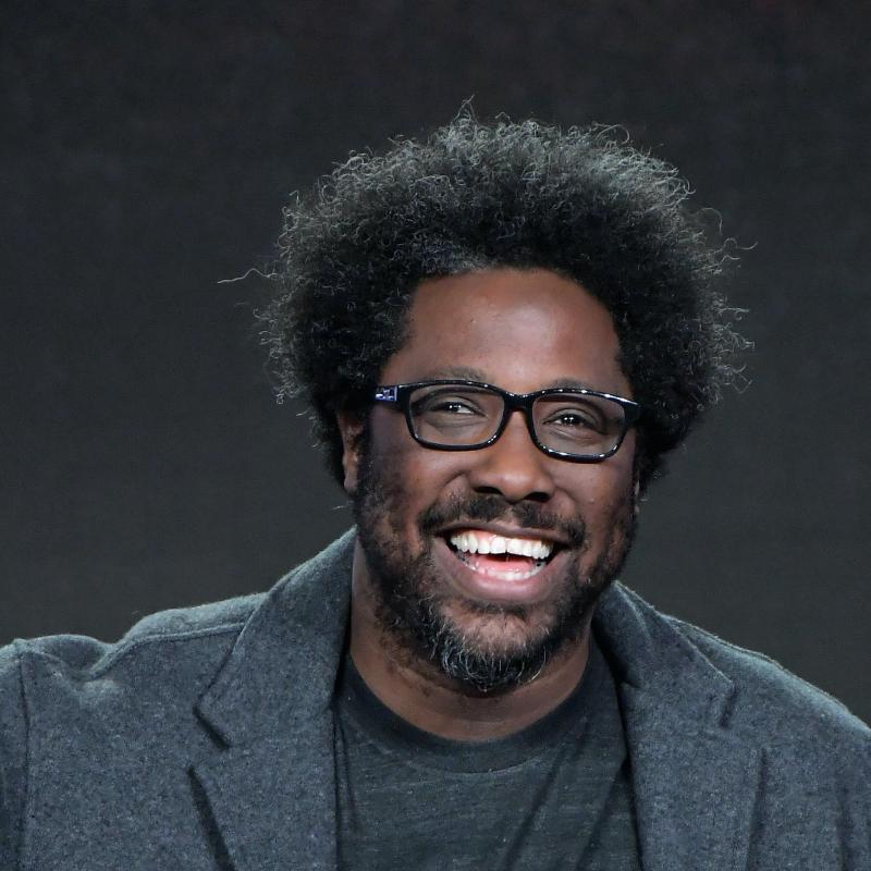 Comedian W. Kamau Bell laughs on stage against a black backdrop